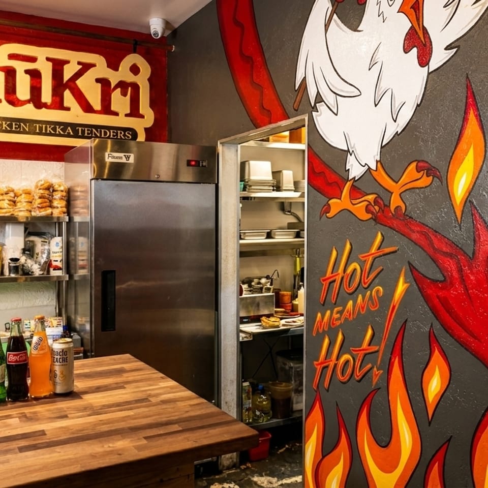 Interior of Kukri Chicken Tikka Tenders restaurant with a counter, shelves of food, and a fiery chicken mural.