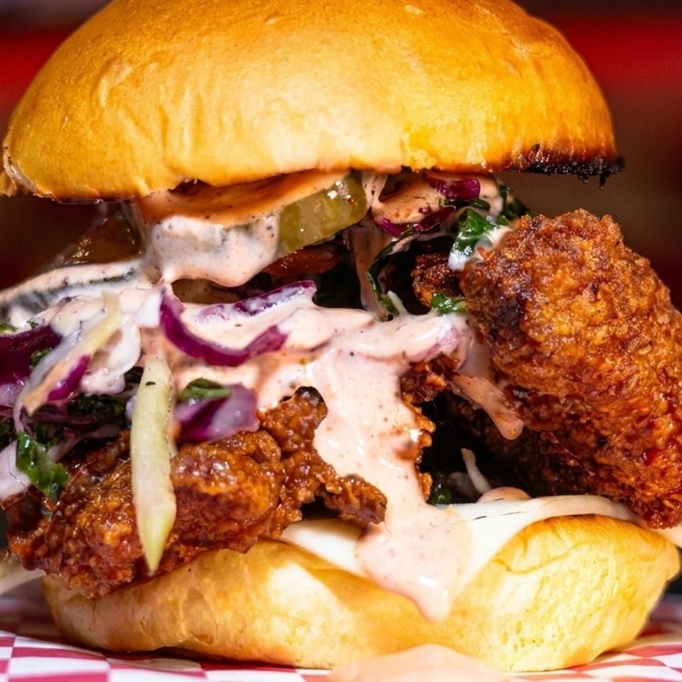 Close-up of a fried chicken sandwich with coleslaw, pickles, and sauce on a brioche bun.