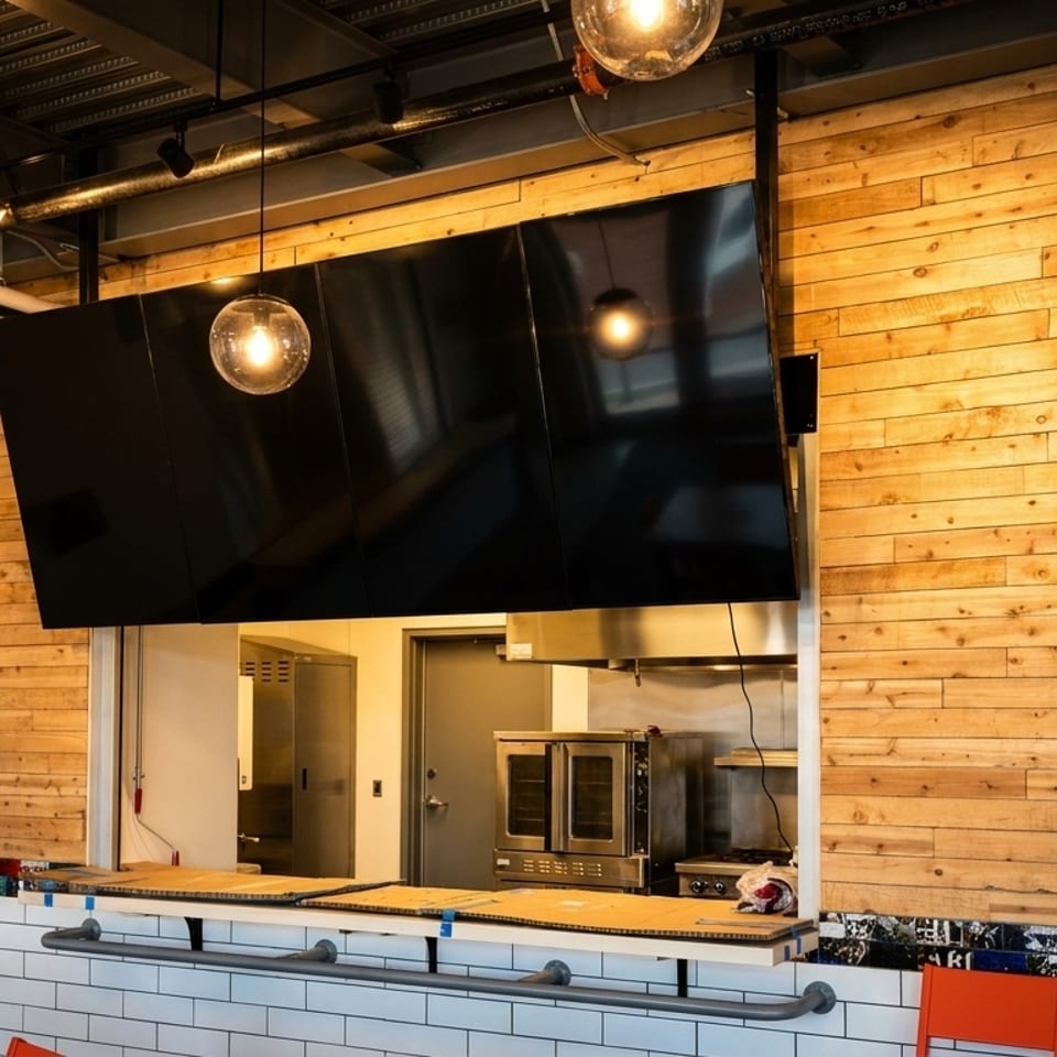 A modern restaurant interior with a wooden wall, large black screens, and an open kitchen pass-through.